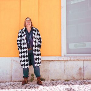 Ginny Evans-Pollard stands smiling against a vibrant orange wall, wearing a bold black-and-white patterned coat, glasses, jeans, and brown boots. The image reflects authenticity, confidence, and individuality, celebrating the idea that professionalism and self-expression can coexist.